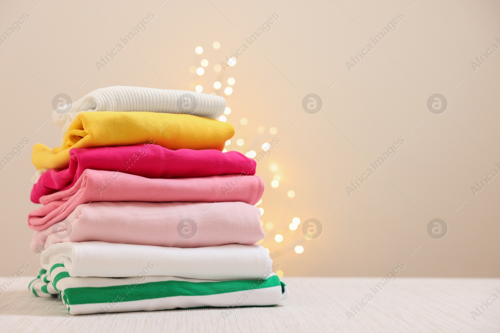 Stack of folded clothes on wooden table against beige background with blurred lights, space for text Photo of Stack of folded clothes on wooden table against beige background with blurred lights, space for text