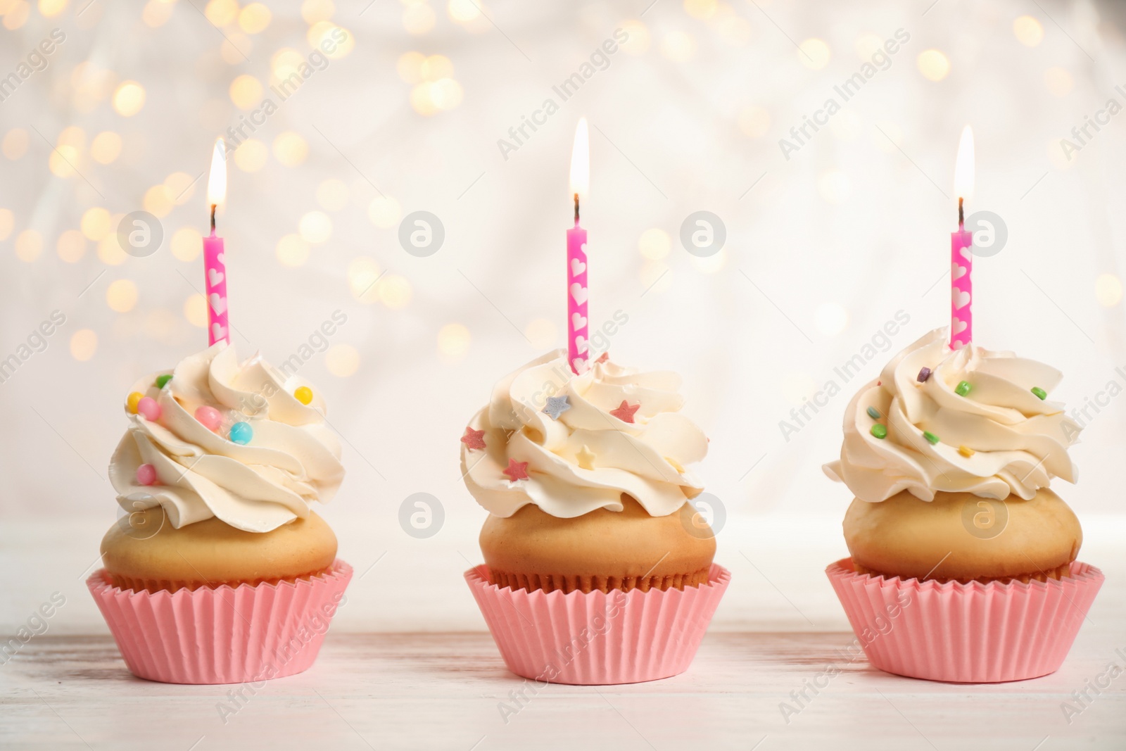 Photo of Birthday cupcakes with candles on white wooden table against blurred lights