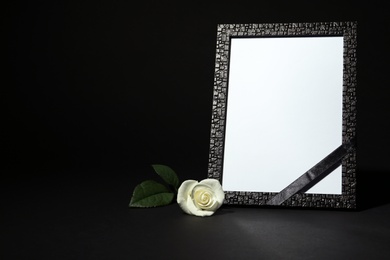 Photo of Funeral photo frame with ribbon and white rose on dark table against black background. Space for design