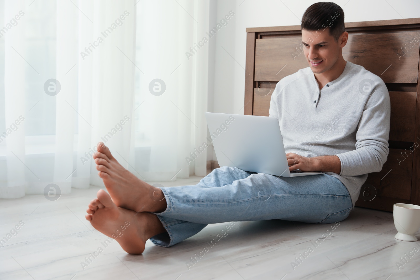 Photo of Man with cup of drink and laptop sitting on warm floor at home. Heating system