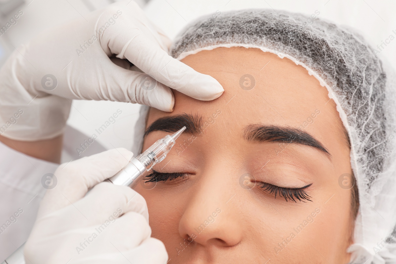 Photo of Young woman undergoing procedure of permanent eyebrow makeup in tattoo salon, closeup