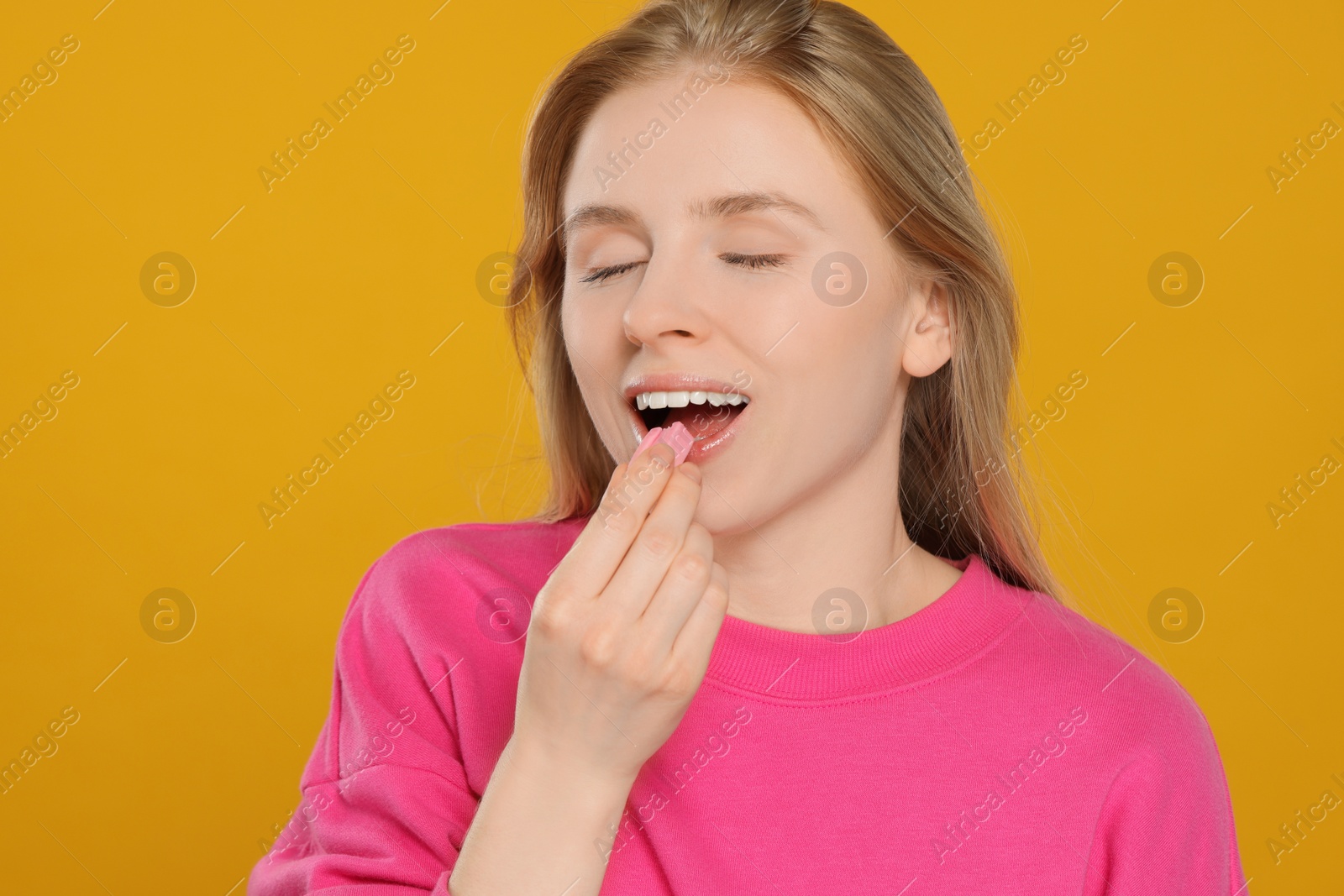 Photo of Happy young woman putting chewing gum into mouth on yellow background