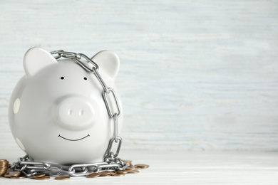 Photo of Piggy bank with steel chain and coins on white wooden table, space for text. Money safety concept