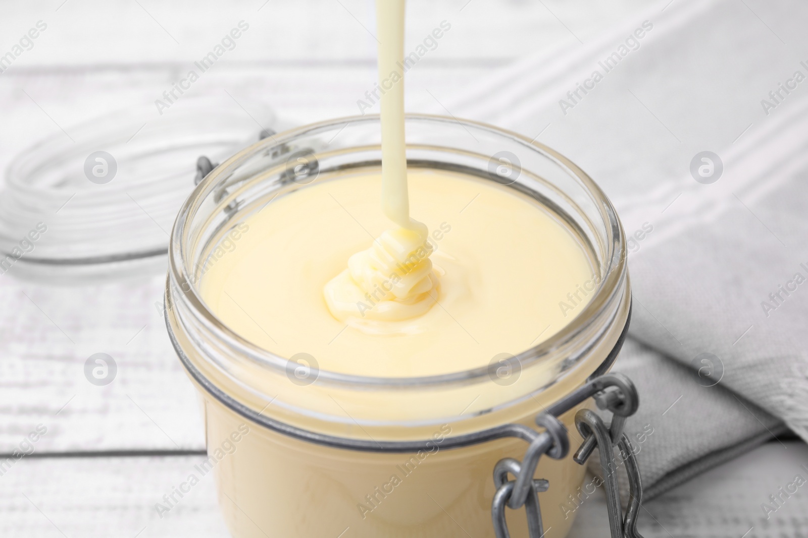 Photo of Pouring tasty condensed milk into jar on white table, closeup
