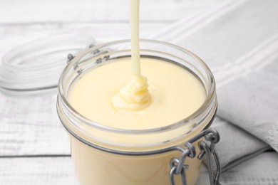 Photo of Pouring tasty condensed milk into jar on white table, closeup