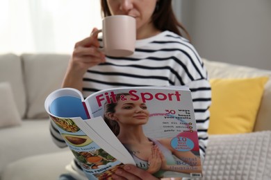 Woman with cup of drink reading sports magazine at home, closeup