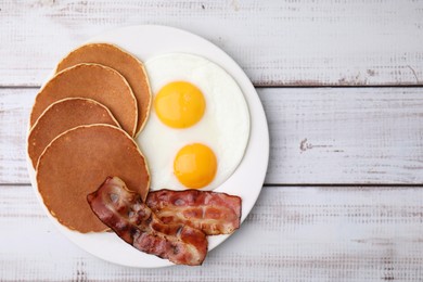 Tasty pancakes with fried eggs and bacon on white wooden table, top view. Space for text