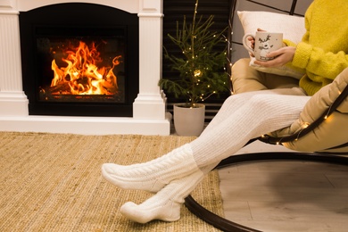 Woman with cup of drink sitting near burning fireplace at home, closeup