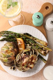 Photo of Tasty grilled artichokes and asparagus served on beige textured table, flat lay