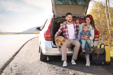 Photo of Parents, their daughter and dog sitting in car trunk near road, space for text. Family traveling with pet