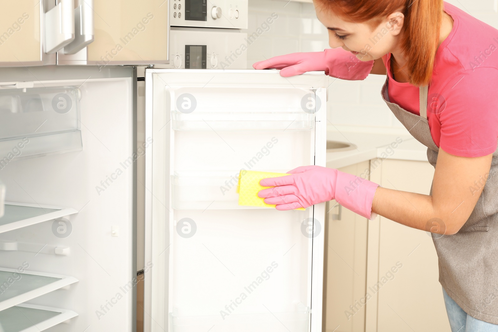 Photo of Woman in protective gloves cleaning refrigerator with rag indoors