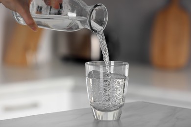 Woman pouring water from bottle into glass at light grey marble table in kitchen, closeup