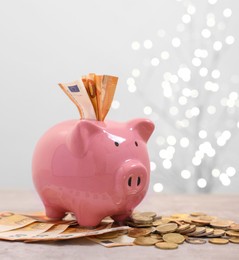 Photo of Piggy bank with euro banknotes and coins on grey table against blurred lights, space for text