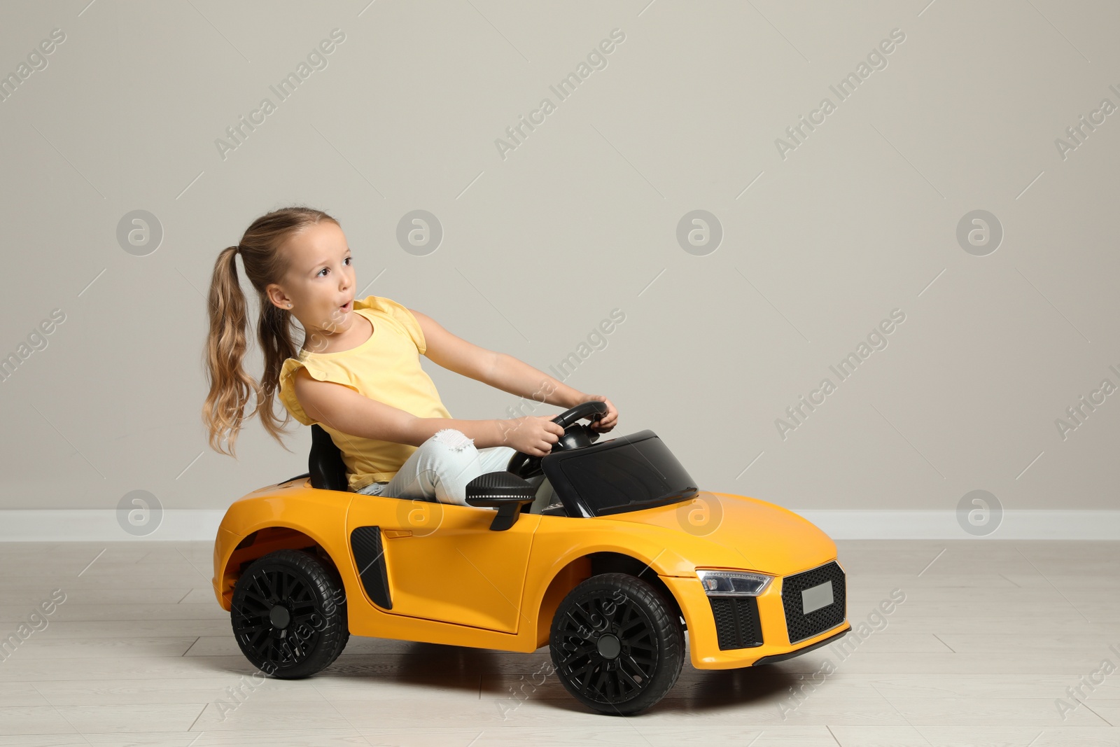 Photo of Cute little girl driving children's electric toy car near grey wall indoors. Space for text