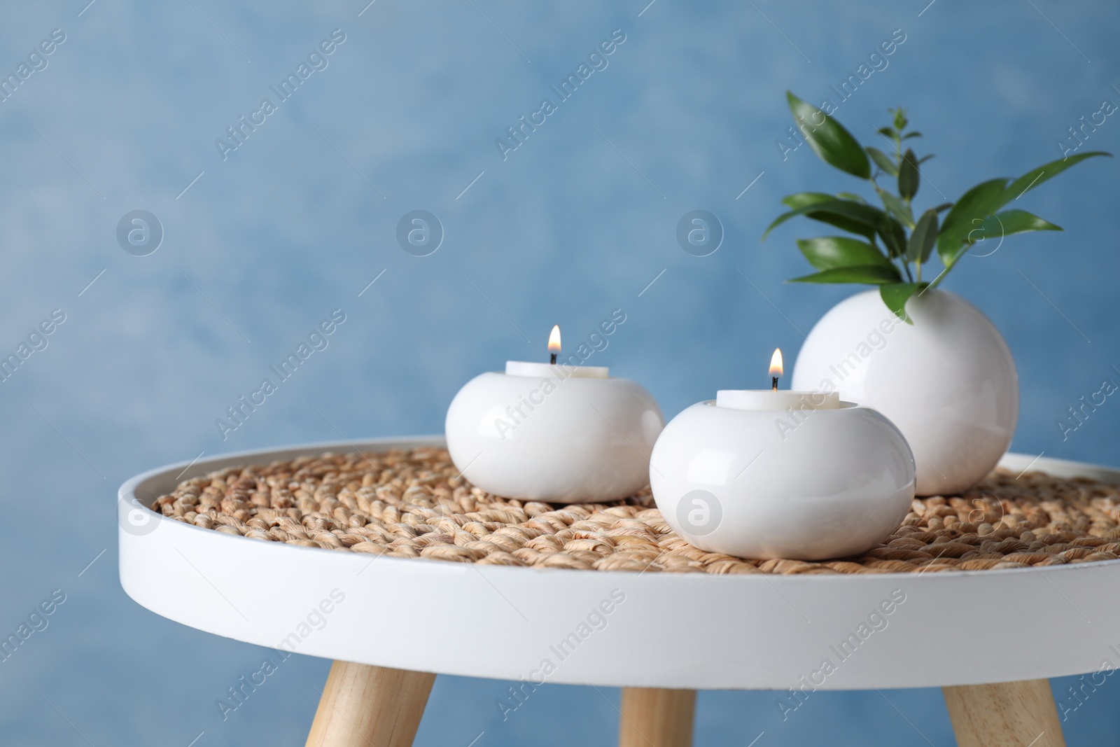 Photo of Burning candles and plant on white table against light blue background, space for text