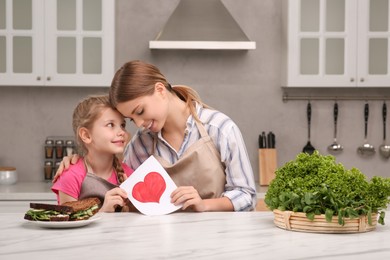 Photo of Little daughter congratulating mom with greeting card in kitchen. Happy Mother's Day