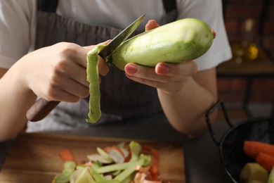 Photo of Woman peeling fresh zucchini with knife at table indoors, closeup