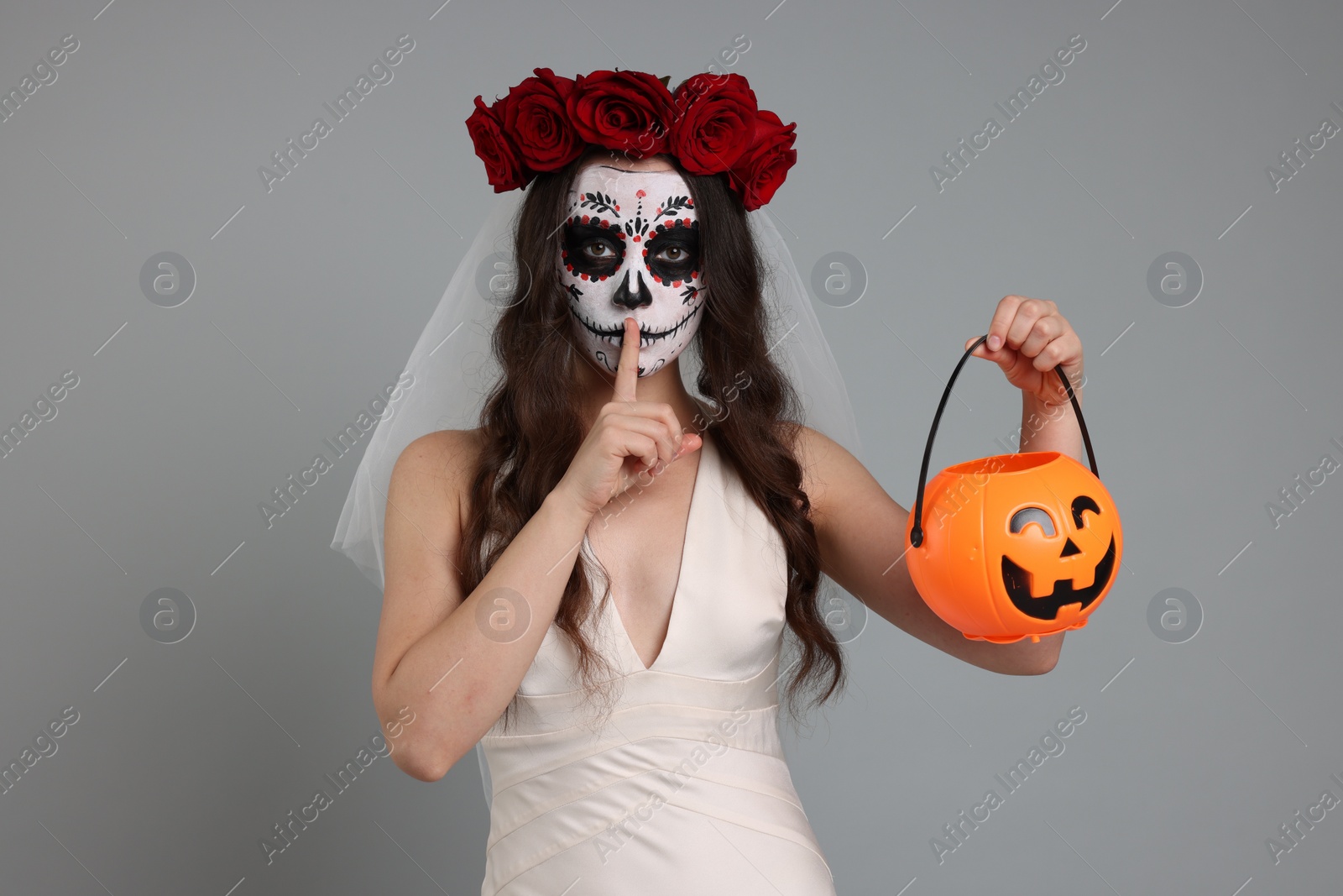 Photo of Young woman in scary bride costume with sugar skull makeup, flower crown and pumpkin bucket showing shush gesture on light grey background. Halloween celebration