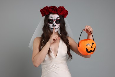 Young woman in scary bride costume with sugar skull makeup, flower crown and pumpkin bucket showing shush gesture on light grey background. Halloween celebration