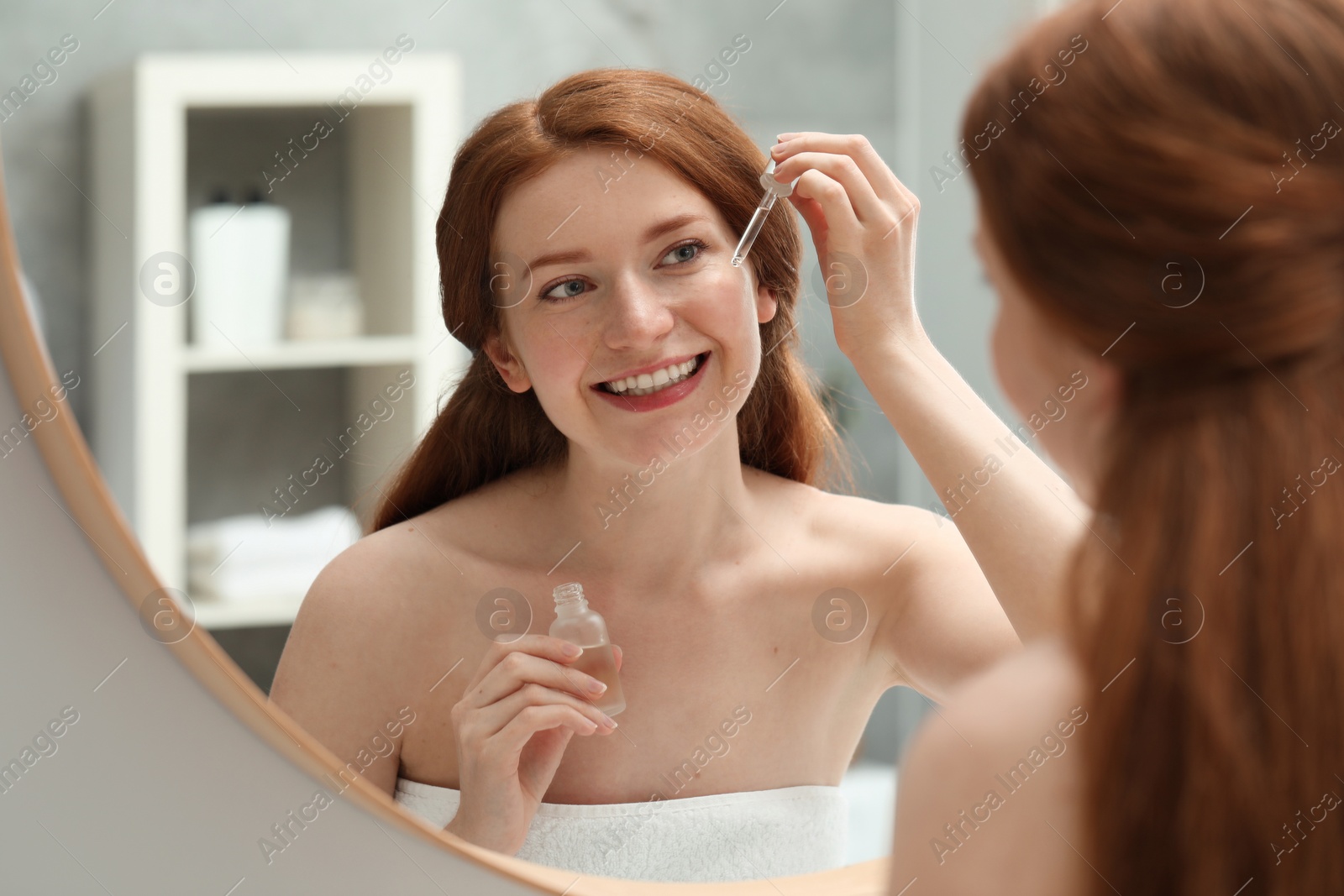 Photo of Smiling woman with freckles applying cosmetic serum onto her face near mirror in bathroom