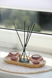 Photo of Reed diffuser with burning candles and wooden tray on white window sill