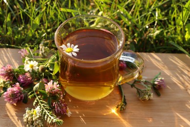 Cup of aromatic herbal tea and different wildflowers on green grass outdoors, closeup