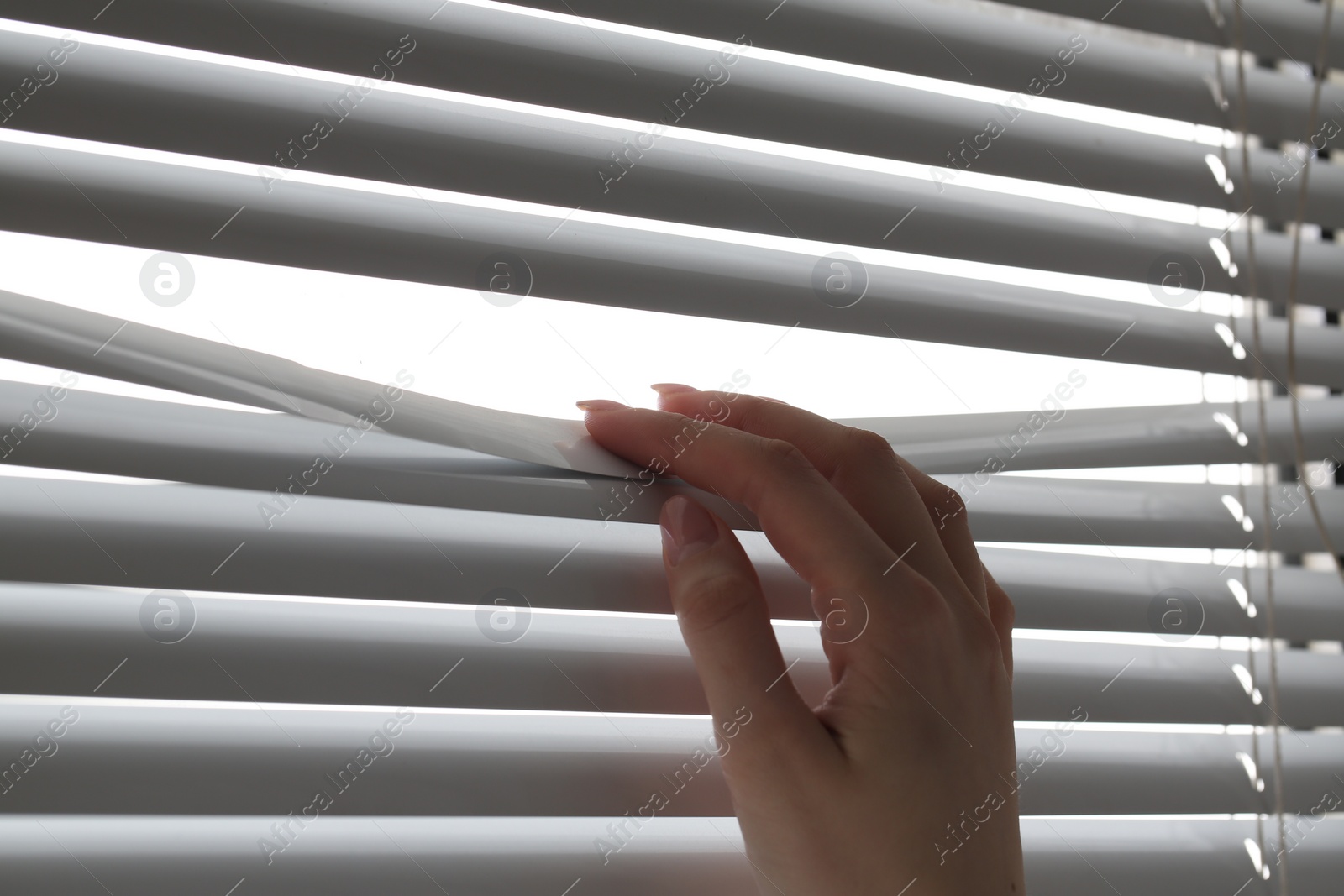 Photo of Woman separating slats of white blinds indoors, closeup