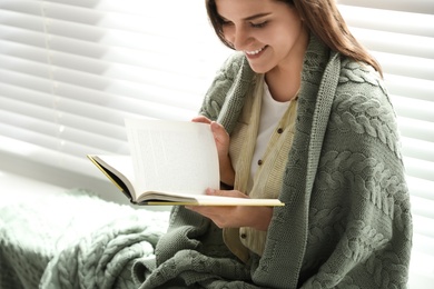 Woman covered with warm green plaid reading book near window indoors