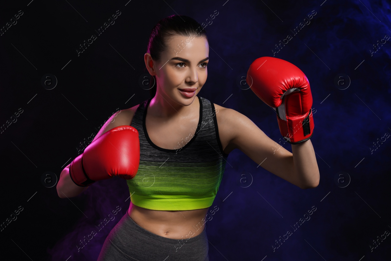 Photo of Portrait of beautiful woman wearing boxing gloves training in color lights on black background