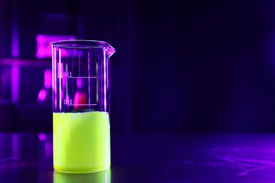 Photo of Laboratory beaker with luminous liquid on table against dark background, space for text
