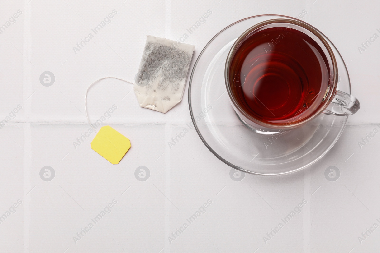 Photo of Tea bag and cup of aromatic drink on white tiled table, top view. Space for text