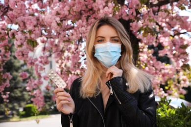 Photo of Woman with pills and protective mask near blossoming tree outdoors. Seasonal pollen allergy