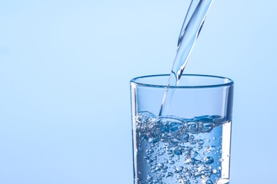 Pouring water into glass against light blue background, closeup. Space for text