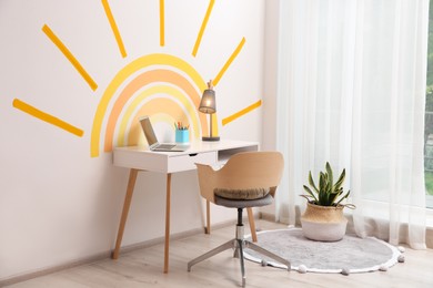 Photo of Laptop, stationery and lamp on white table near wall with painted sun indoors. Interior design
