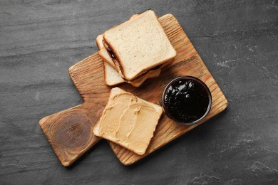 Photo of Tasty peanut butter sandwiches and jam on dark gray table, top view