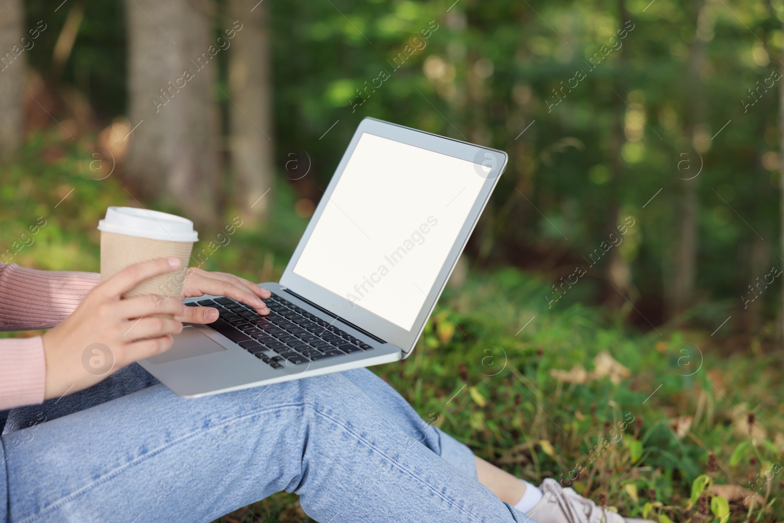 Photo of Young woman with cup of coffee working on laptop in forest, closeup