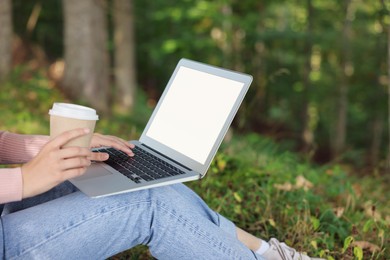Photo of Young woman with cup of coffee working on laptop in forest, closeup