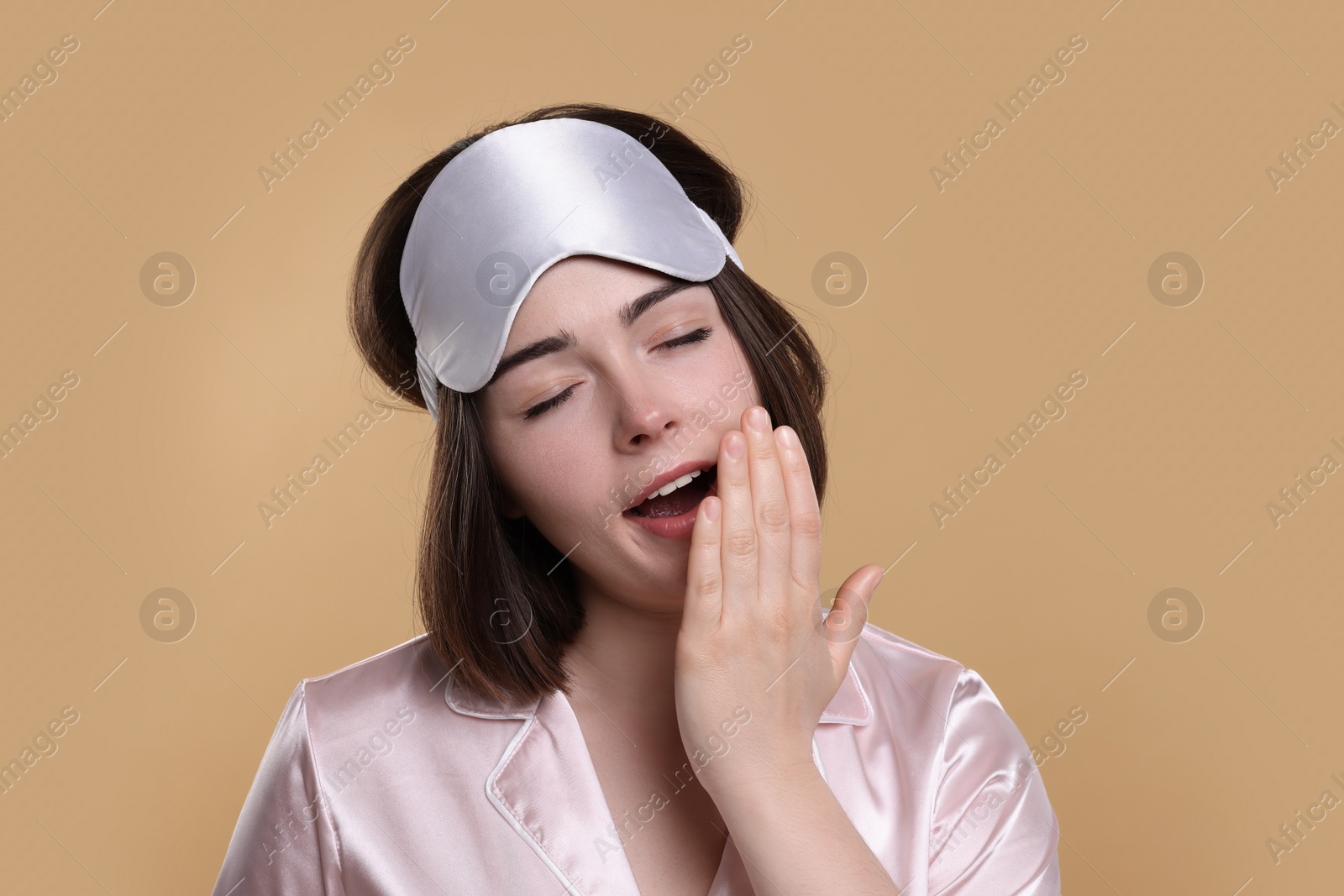 Photo of Tired young woman with sleep mask yawning on beige background. Insomnia problem
