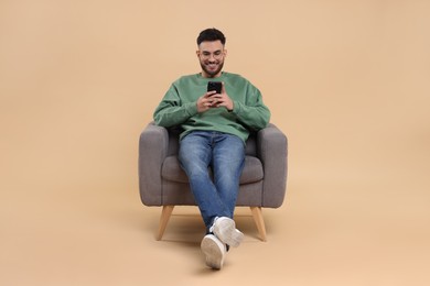 Photo of Happy young man using smartphone in armchair on beige background