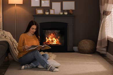 Beautiful young woman reading magazine on floor near fireplace at home. Space for text