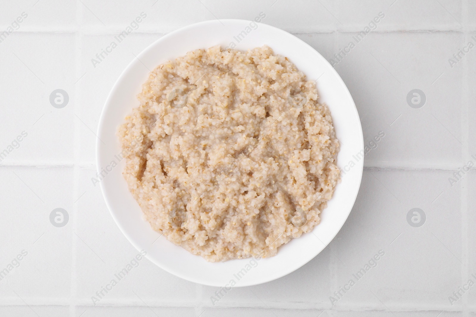 Photo of Delicious barley porridge in bowl on white tiled table, top view