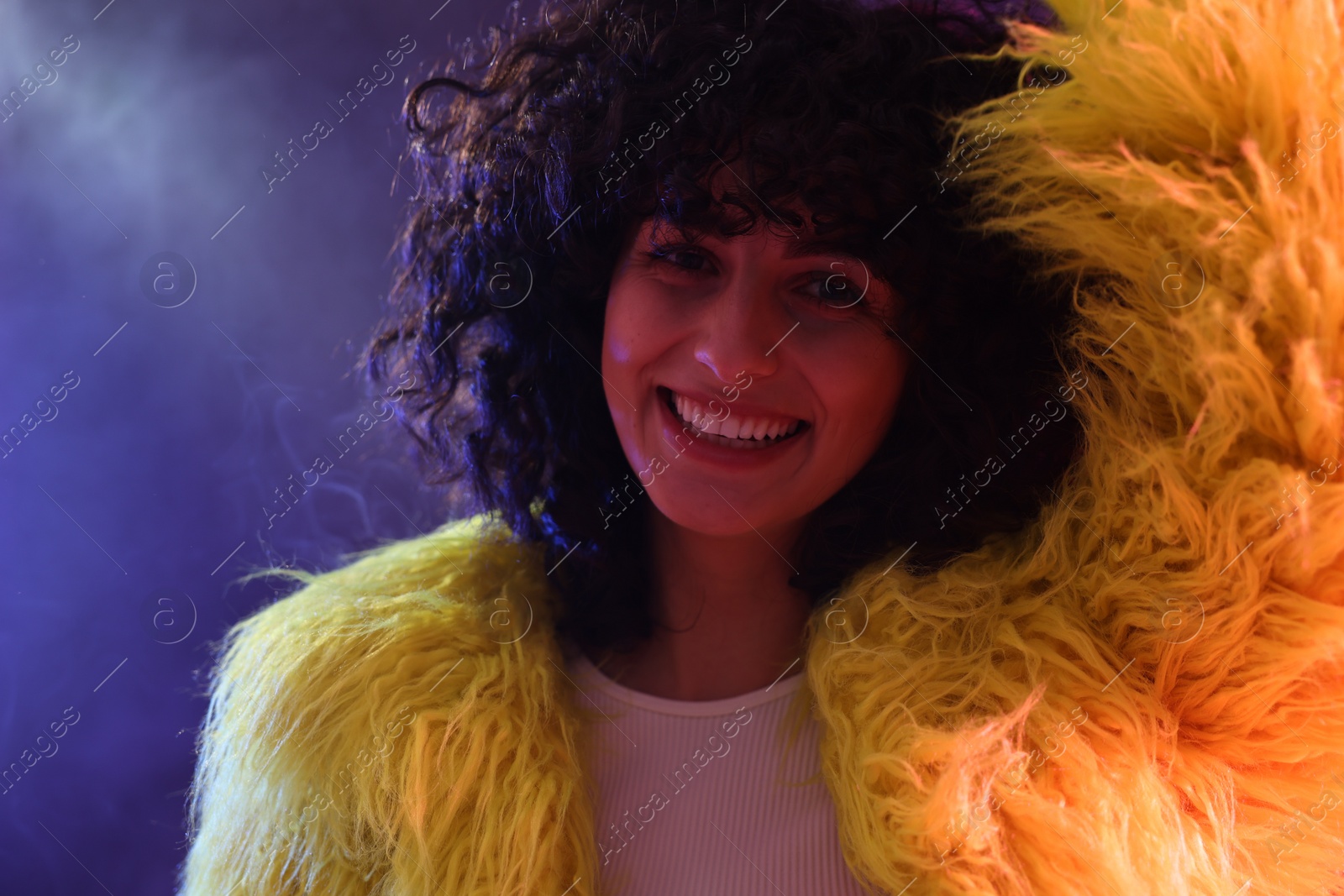 Photo of Beautiful young woman in yellow fur coat on color background in neon lights and smoke