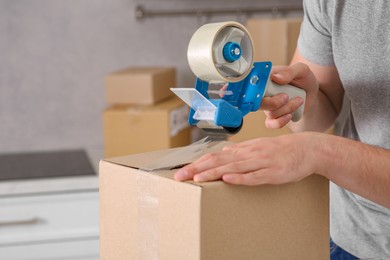 Photo of Man taping box with adhesive tape dispenser in kitchen, closeup