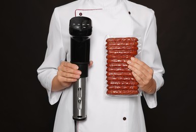 Photo of Chef holding sous vide cooker and sausages in vacuum pack on black background, closeup