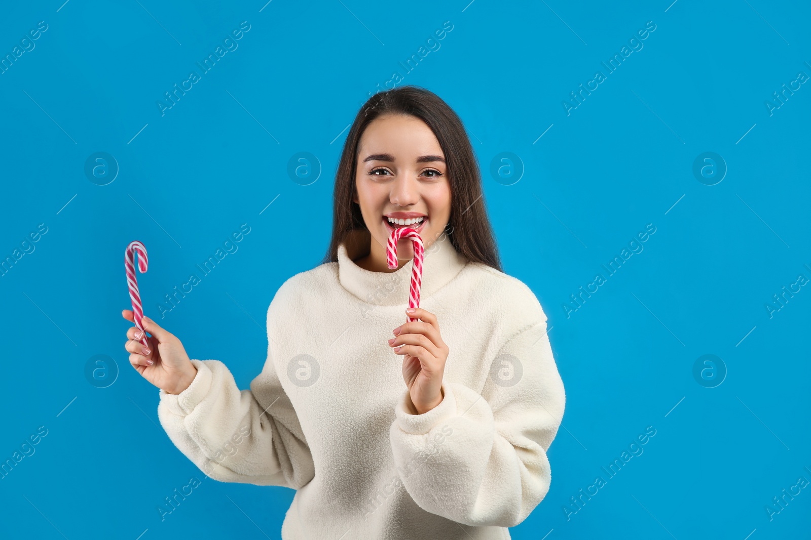 Photo of Young woman in beige sweater holding candy canes on blue background. Celebrating Christmas