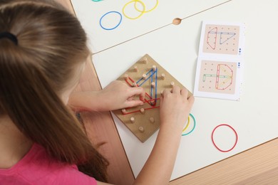 Photo of Motor skills development. Girl playing with geoboard and rubber bands at white table, above view