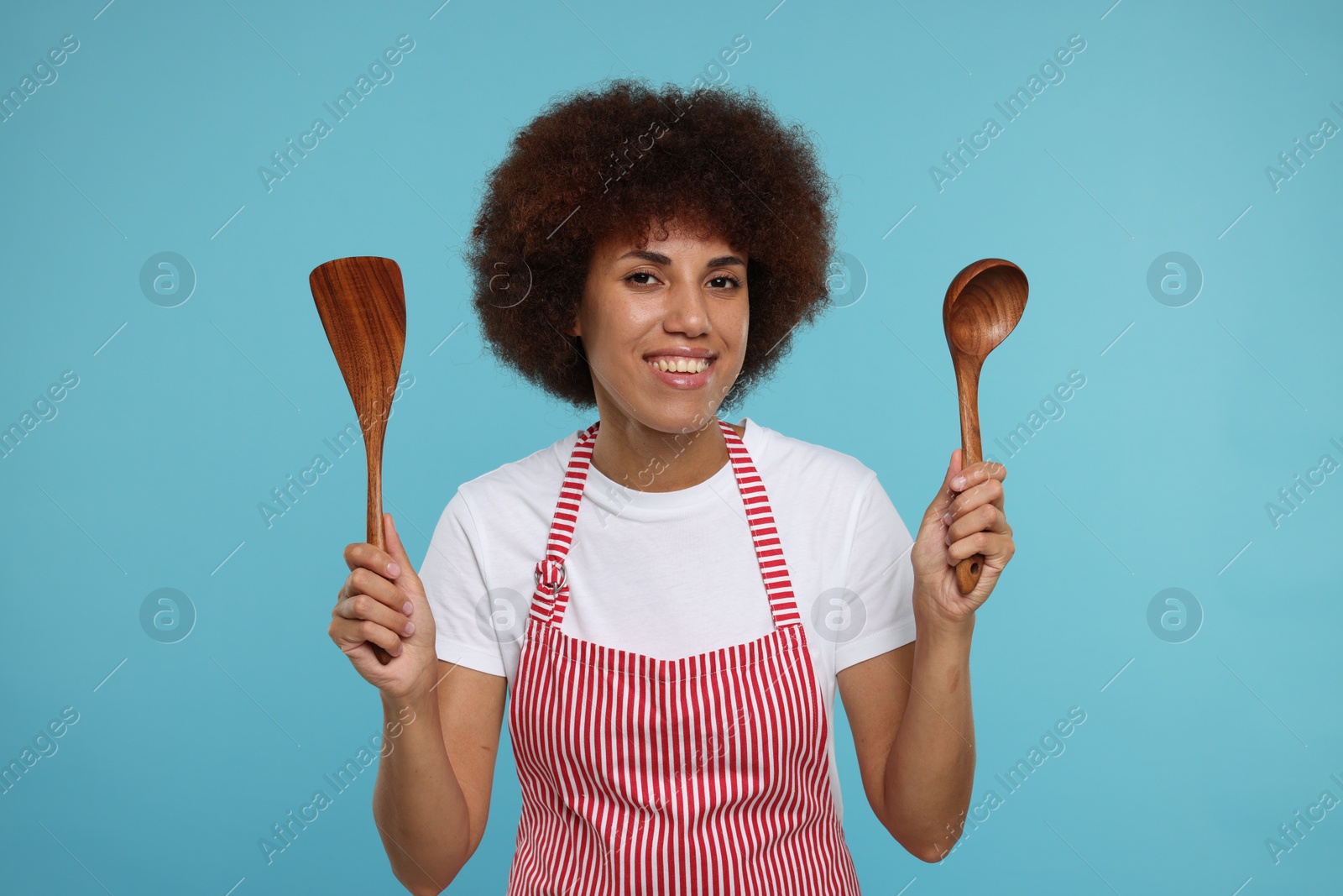 Photo of Happy young woman in apron holding spoon and spatula on light blue background