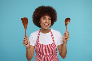 Happy young woman in apron holding spoon and spatula on light blue background