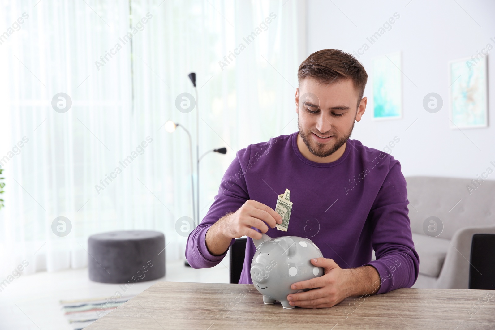 Photo of Young man putting money into piggy bank at table indoors. Space for text