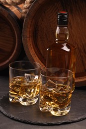 Photo of Whiskey with ice cubes in glasses, bottle and wooden barrels on black table